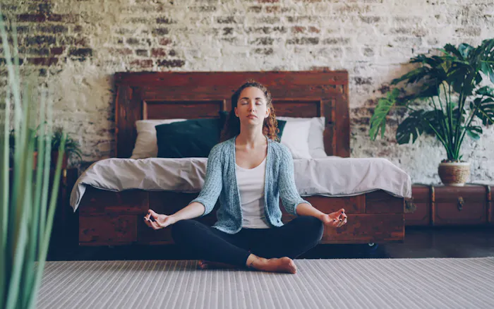 A person sitting cross-legged on the floor of a quiet room, eyes closed, in a calm meditative posture