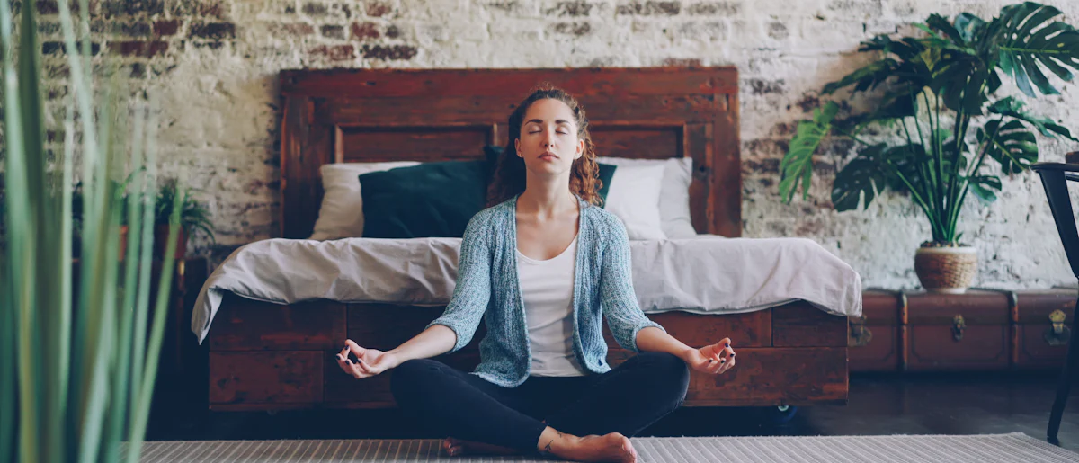 A person sitting cross-legged on the floor of a quiet room, eyes closed, in a calm meditative posture